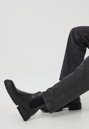 Czarne skórzane botki Chelsea z elastycznymi panelami bocznymi i grubymi gumowymi podeszwami. Noszone z ciemnoszarymi spodniami slim fit na jasnym tle.