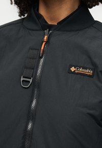 Κοντινό πλάνο μαύρου μπουφάν Columbia Sportswear με εμπρόσθιο φερμουάρ, πορτοκαλί κεφαλή φερμουάρ και μαύρο ιμάντα με μεταλλικό κλιπ στο στήθος.