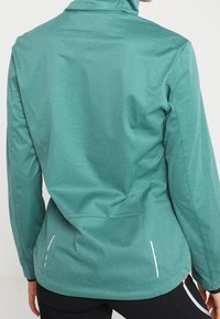 Tealtkleurig sportjacket met een hoge kraag, een gladde textuur en reflecterende accenten langs de zijnaad. Licht getailleerd ontwerp voor bewegingsvrijheid.