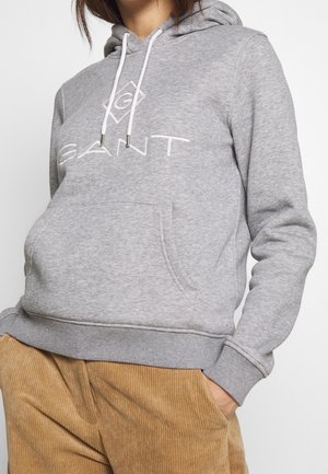 Persoon draagt een grijze GANT hoodie met een voorzak en witte trekkoorden, gecombineerd met bruine corduroy broeken, handen in de zakken.