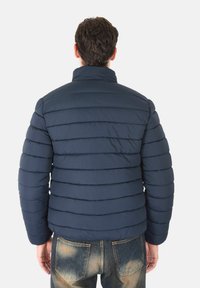 Giacca trapuntata blu navy con cuciture orizzontali, colletto alto e polsini elasticizzati, abbinata a jeans chiari con aree sbiadite.