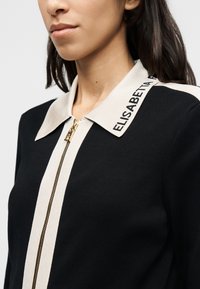 Μαύρο πανωφόρι με φερμουάρ και μεγάλο λευκό γιακά που έχει κεντημένο κείμενο. Το ύφασμα έχει ριμπ υφή και χρυσό φερμουάρ.