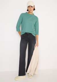 Top in teal chiaro con texture e maniche a tre quarti, abbinato a pantaloni in denim scuri a zampa d'elefante. Cappello beige e giacca beige chiaro appoggiati al braccio.