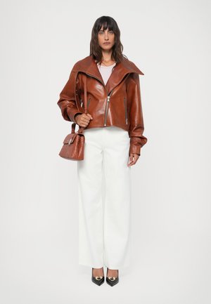 Veste en cuir marron avec un large col, fermeture éclair et manches longues. Associée à un pantalon large blanc et à un petit sac à main texturé.