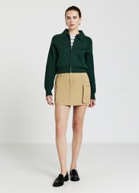 Maglione verde a maglia con zip e colletto, abbinato a una gonna mini in cargo beige con tasche laterali e scarpe nere flat.