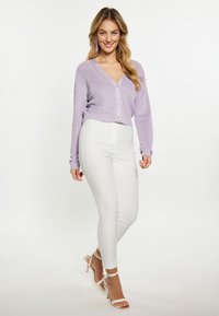 Cardigan cropped lilla con scollatura a V e chiusura con bottoni, abbinato a pantaloni skinny bianchi a vita alta e tacchi beige con cinturini.