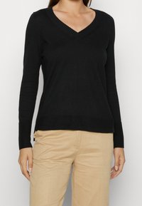 Svart långärmad v-ringad tröja med ribbad krage och muddar, tillverkad av mjukt stickat material, bärs över beige byxor.