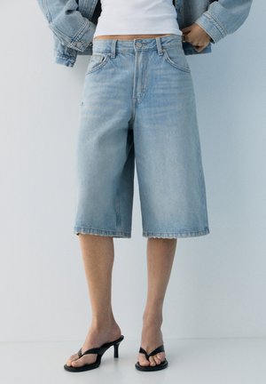 Person iført lyseblå denim shorts til knæene, hvid top, denimjakke og sorte sandaler med hæl, stående mod en hvid væg.