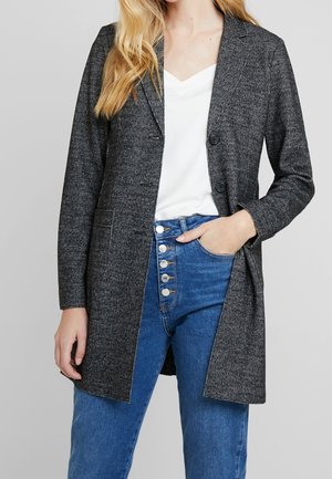 Femme portant un long blazer gris charbon sur un haut blanc et un jean taille haute bleu à cinq boutons, main dans la poche.