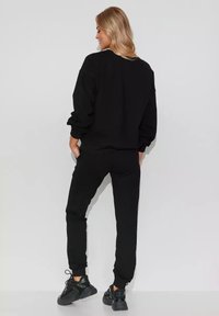 Czarny zestaw bluzy z długim rękawem i dresów wykonany z miękkiego materiału. Posiada ściągacze, luźny krój i jest zestawiony z czarnymi butami sportowymi.