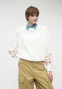 Baltā kapučjaka ar sarkaniem sirds rakstiem uz piedurknēm, ar apaļu kakla izgriezumu un brīvu piegriezumu. Apakšā valkāta gaiši zila džinsa kreklu.