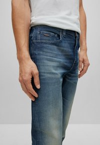 Jeans de mezclilla azul oscuro con un ligero degradado, que cuentan con cinco bolsillos, un lazo para cinturón y una etiqueta en el bolsillo delantero. Textura suave.