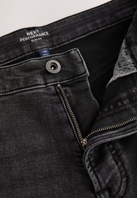 Jeans nero slim fit con cerniera parzialmente aperta, bottone visibile con testo ed etichetta della taglia 34R all'interno del cinturino.