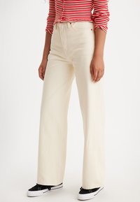 Pantalons large couleur crème avec une taille haute, en denim. Associés à des baskets noires, ils présentent des coutures discrètes et des poches.