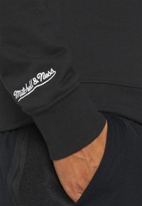 Handen i fickan, iförd svart sweatshirt med vitt broderat "Mitchell & Ness"-logotyp på ärmavslut.