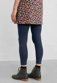 Tamno plave tajice u kombinaciji s tunikom s uzorkom koja prikazuje višebojne listove. Model nosi sive gležnjače s drvenim potpeticama.