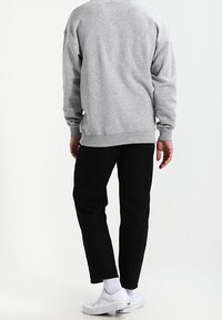Sudadera gris con puños acanalados, llevada sobre pantalones negros. Se ven zapatillas blancas y calcetines blancos. El fondo es liso y de color claro.