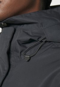 Gros plan sur le col d'une veste noire avec une fermeture éclair et un cordon de serrage ajustable sur une peau foncée.