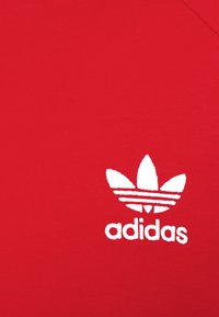 Camiseta de algodón roja con un logo de Adidas en blanco que presenta tres franjas en forma de hoja y el nombre de la marca en negrita debajo del logo. Textura suave.