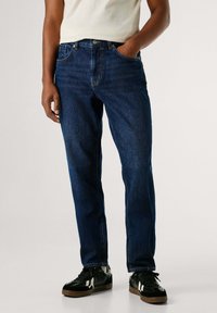 Vaqueros de mezclilla azul oscuro con corte recto. Cuenta con un diseño clásico de cinco bolsillos y cierre de botón metálico. Llevados con zapatillas negras.