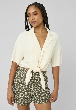 Femme aux cheveux tressés portant un chemisier court crème à manches courtes noué à la taille et un short vert à motifs floraux, debout devant un fond uni.