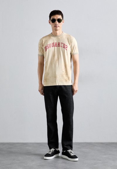 Beige T-Shirt mit rotem "DSQUARED2"-Text, ausgestattet mit kurzen Ärmeln und sichtbaren Schmutzflecken. Kombiniert mit dunklen Jeans und schwarzen Sneakern.