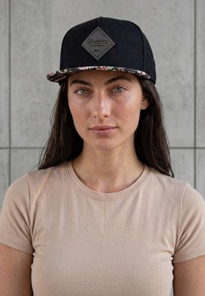 Nuori nainen, jolla on pitkät tummat hiukset, musta lippalakki jossa on kukallinen lippa ja beige t-paita, seisoo harmaan laatoitetun seinän edessä.