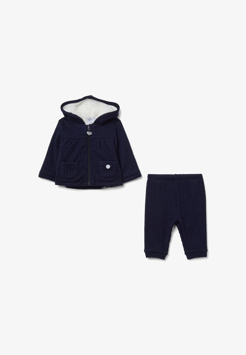 Felpa a collo alto blu navy con fodera in pelliccia e tasche frontali, abbinata a pantaloni in maglia coordinati. Chiusura con zip e dettaglio a cerchio.