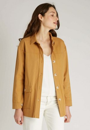 Veste jaune moutarde en coton, dotée d'une fermeture à boutons, de deux poches avant et d'un col classique. Le tissu a une texture lisse.