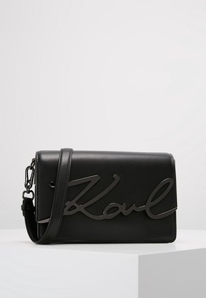 Sac à bandoulière en cuir noir avec un grand logo stylisé "Karl" à l'avant et une sangle réglable avec boucle métallique.