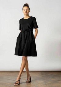 Schwarzes, kurzärmeliges Kleid mit tailliertem Schnitt, seitlichen Taschen und einem fließenden Rock. Hergestellt aus weichem Stoff, mit rundem Ausschnitt und Gürtel zum Binden.