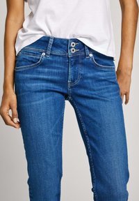 Vaqueros de mezclilla azul con un diseño ajustado, cierre de botón doble y costuras de contraste claras. Incluyen bolsillos frontales y una textura suave.