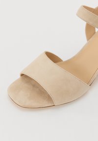 Beige suede sandal featuring a wide front strap and ankle strap, with a minimalist design and open toe outlining.