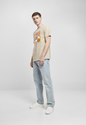 Beige grafisk t-skjorte med en animert karakter som holder basketballer, matchet med lyseblå jeans og hvite sneakers, stående på en enkel bakgrunn.