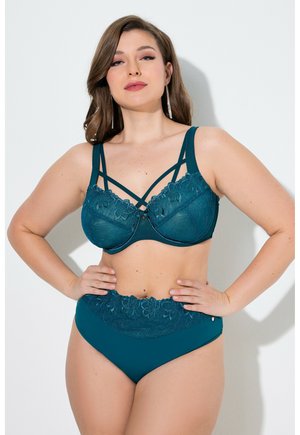 Vrouw poseert in een tealkleurige kanten lingerieset met een beha met bandjes en hoge taille slipjes met bloemrijke kantdetails, handen op de heupen tegen een effen achtergrond.
