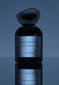 Czarna szklana butelka perfum o zaokrąglonym kształcie, z błyszczącą nakrętką przypominającą kamień oraz srebrną etykietą z tekstem. Odbita na ciemnej powierzchni.