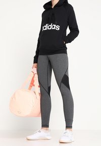 Schwarzer Hoodie mit weißem Logo, graue Leggings mit schwarzen Akzenten, rosa Sporttasche und weiße Sportschuhe. Glatter Stoff, figurbetonter Schnitt.
