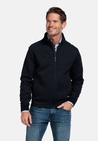 Zwarte zip-up jack met groene accenten op de schouders, geribde manchetten en een zachte textuur. Gedragen over een gestreept shirt, stijlvol met blauwe spijkerbroeken.