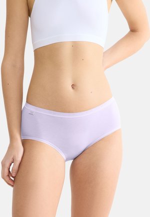 Frau trägt hellviolette Baumwollunterwäsche und ein weißes Sport-BH, steht mit einer Hand auf der Hüfte vor einem einfarbigen Hintergrund.