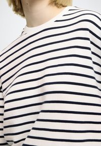 Bawełniana koszulka z granatowymi poziomymi paskami na jasnym tle. Posiada prążkowany kołnierz i luźny krój. Gładka faktura.