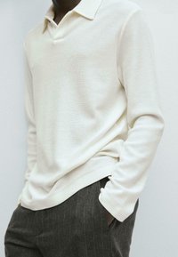 Polo a maniche lunghe di colore crema realizzato in tessuto testurizzato, con colletto classico e vestibilità rilassata, abbinato a pantaloni scuri a righe sottili.