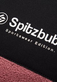Tessuto nero con scritte bianche che recitano "Spitzbub Sportswear Edition." Un materiale rosa testurizzato è visibile lungo il bordo inferiore.