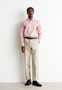 Camicia rosa chiaro con bottoni, pantaloni beige strutturati, cintura nera e scarpe nere. Il modello si trova davanti a uno sfondo bianco semplice.