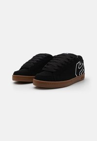 Schwarze Wildledersneakers mit einer braunen Gummisohle, ausgestattet mit schwarzen Schnürsenkeln und einem weißen Logodetail an der Seite. Glatte Textur insgesamt.