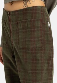 Pantalons en velours côtelé vert avec un motif à carreaux en vert foncé et bordeaux, présentant une fermeture éclair à la taille et deux poches avant.