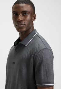 Jaunas vyras dėvi tekstūruotą tamsiai pilką polo marškinėlį su baltu dryžiu ant apykaklės ir rankovės, stovintis prie šviesios, paprastos fono.