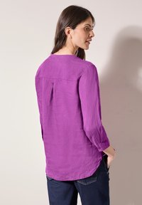 Lila linneblus med rak skärning, trekvartslånga ärmar och detalj med bakveck, i kombination med mörkblå jeans.