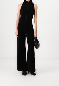 Mono negro con un corpiño ajustado, cuello alto, cremallera completa y piernas anchas. Presenta una tela suave, detalles de costura sutiles y se complementa con un bolso negro.