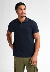 Námořnické polo tričko z texturované látky, se zapínáním na tři knoflíky a kontrastním pruhovaným límcem. Nosené s olivovými cargo šortkami.