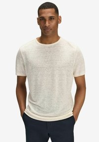 Lys beige kortærmet T-shirt lavet af blødt, struktureret stof. Har en klassisk rund hals og en afslappet pasform. Båret sammen med mørke bukser.
